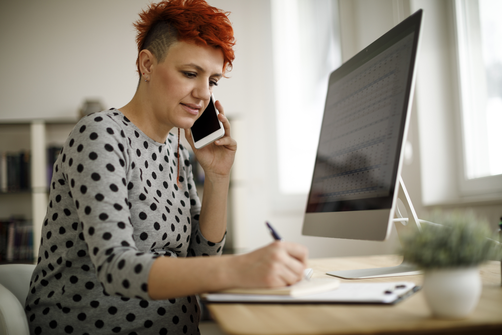 Person with short red hair and a side shave, wearing a polka dot shirt, talking on a smartphone while writing on a notepad at a desk with a large computer monitor. A small plant is on the desk in the foreground.