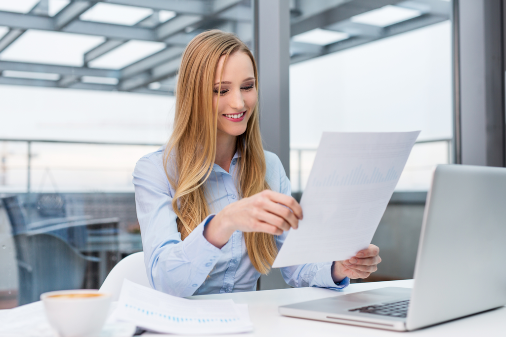 Smiling woman with long blonde hair, wearing a light blue shirt, looking at documents while sitting at a desk with a laptop. A cup of coffee and additional papers are on the desk in the foreground. She is in a bright, modern office with large windows.
