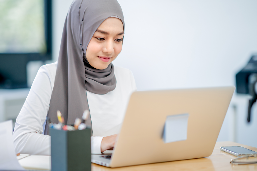 Smiling woman wearing a grey hijab and white shirt, working on a laptop at a desk. A container with pencils and a smartphone are on the desk beside her. The background shows a bright, modern office space.
