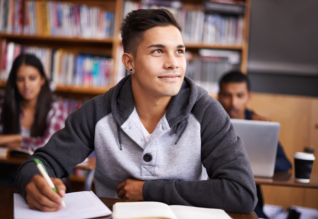 Student in a gray hoodie sitting at a desk in a library or classroom, looking thoughtful while writing in a notebook. Bookshelves filled with books are visible in the background, along with other students studying.