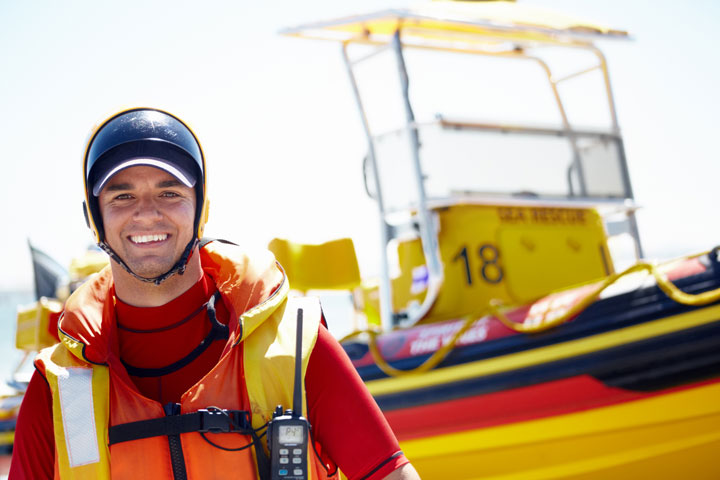 A young, male lifesaver is smiling and wearing a red vest with a yellow lifejacket, a black helmet and a walkie-talkie. He is standing in front of a rescue boat on a beach on a sunny day.