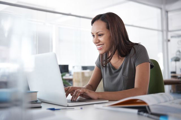A young woman is smiling while working on a laptop in a bright, modern office, with books and papers on the desk.