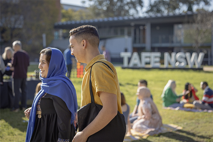 Two smiling students walking outdoors on a sunny day at a TAFE NSW campus. There’s a large TAFE NSW sign and other students sitting on the grass in the background