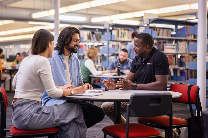 Three people sitting at a round table in a library, engaged in conversation. A TAFE NSW staff member is speaking with two students, with bookshelves and other students in the background.