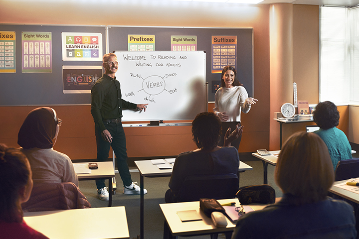 Two teachers are standing in front of the class, smiling and talking to the students. There is a whiteboard with the text:' Welcome to reading and writing for adults' on it