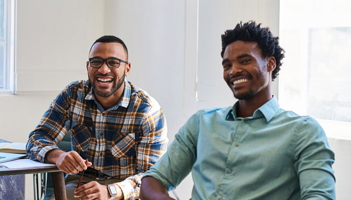 Two young men sit at separate desks, smiling at the camera. The man on the left holds a pen and wears black-rimmed rectangular glasses and a black, white, and red checked shirt. The man on the right wears a light blue button-up shirt.