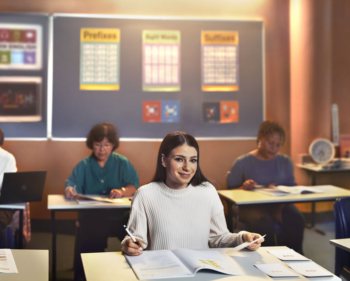 A young woman sits at a desk in a classroom with her study book open and a pen in hand. She is wearing a cream jumper and she is smiling. There are other students working in the background.