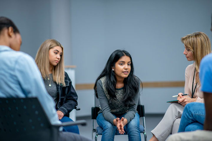 Young people seated in a circle in a room with blue walls, the girl centre leaning forward to speak.