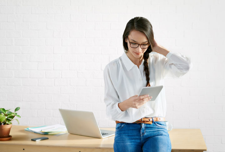 A girl is standing holding a tablet. She is leaning against her desk wearing a white button up shirt tucked into blue jeans.
