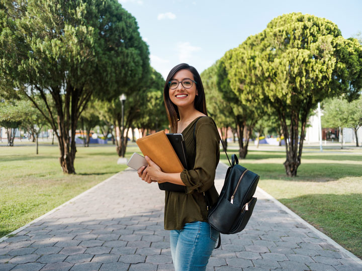 A female student wearing glasses, a green top, jeans and a leather backpack and holding a folder, tablet and mobile phone is standing on a brick path surrounded by grass and trees on a campus and is smiling at the camera.
