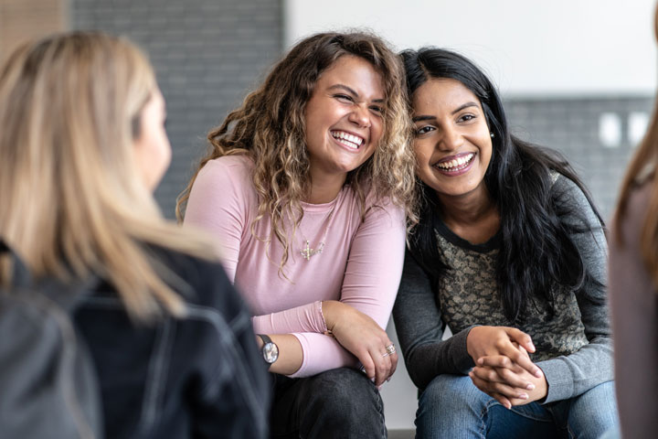 There are two young girls laughing in a group of people. They are also leaning on each other, so they seem to be close friends. The other people have their back to the camera, so they seem to be sitting in a circle chatting.