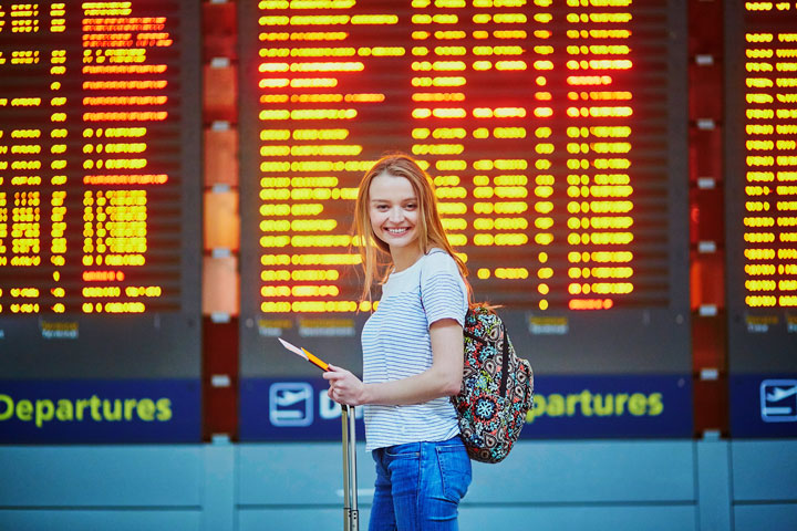 A young female student is standing in front of the flight departure information display in an airport, wearing a backpack, leaning on her luggage and holding her passport and travel documents.