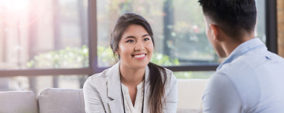 A a young woman seated on a couch by windows and smiling to the young man before her.