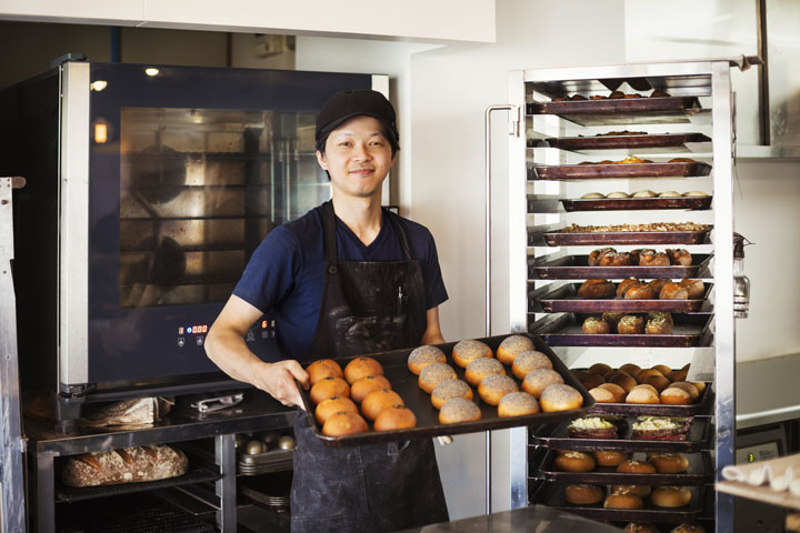 A chef or baker stands in a commercial kitchen, holding a tray of freshly bakes rolls.