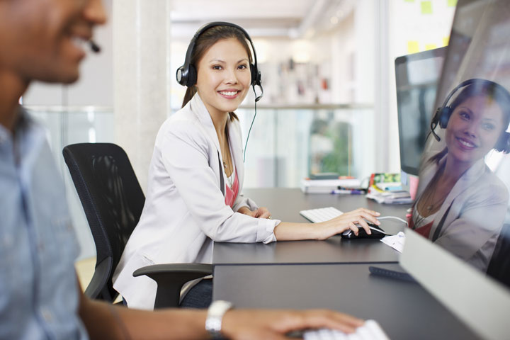 A woman wearing a headset sits at a desk with a computer, smiling at the camera, while a colleague works next to her.