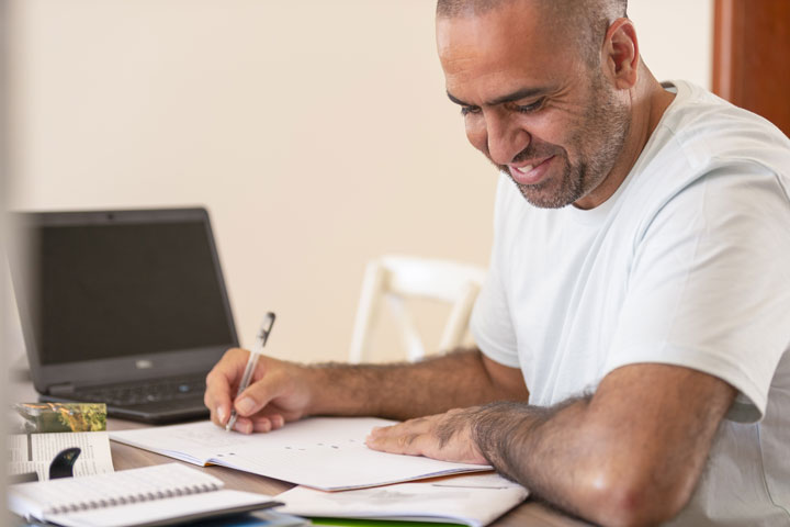 A man sits at his desk, looking down at his notebook. He is holding a pen in his hand and has a laptop on his desk in the background.