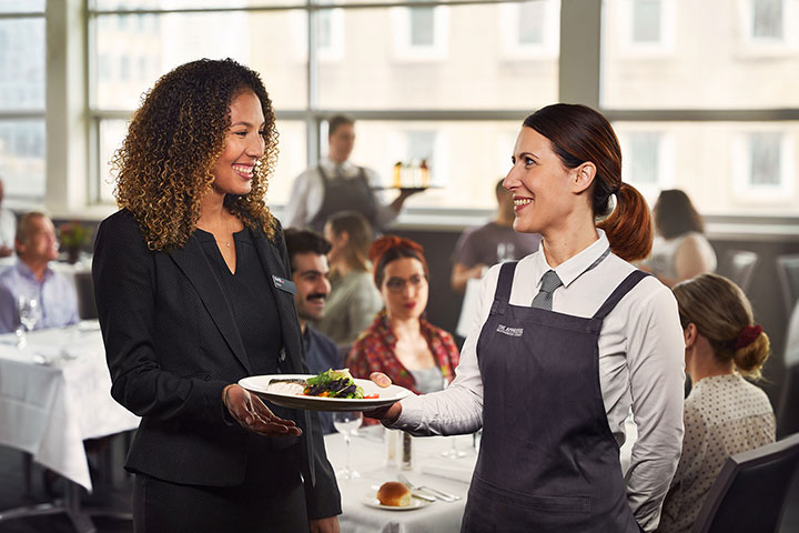 The image shows a professional interaction in a restaurant setting. A smiling server in a uniform, holding a plate of food, is handing it to a woman in business attire. The two appear to be exchanging polite conversation. In the background, diners are seated at tables, engaged in their meals, while other staff members work.