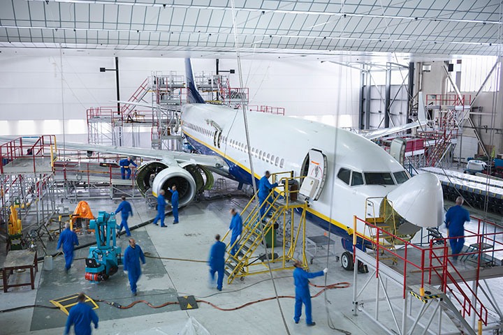 The image shows a large plane in a warehouse that is being services by people in blue uniforms. The workers are slightly blurred, indicating that they are moving around and working on the plane.