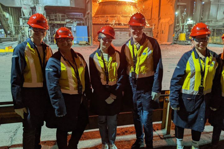 The image shows five people posing for a photo in a steel manufacturing facility. They are wearing protective clothing including long pants, Hi-Vis jackets, Hard Hats and sunglasses. They look happy to be there and to be learning.