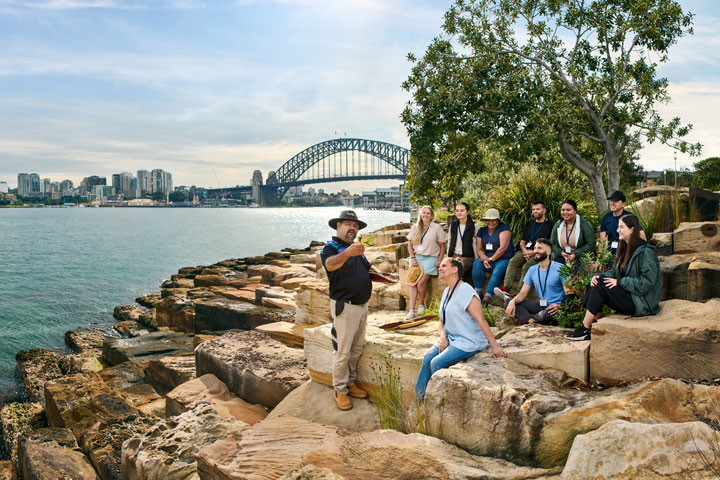 Tour group seated on rocks by Sydney Harbour, listening to a local Indigenous guide.