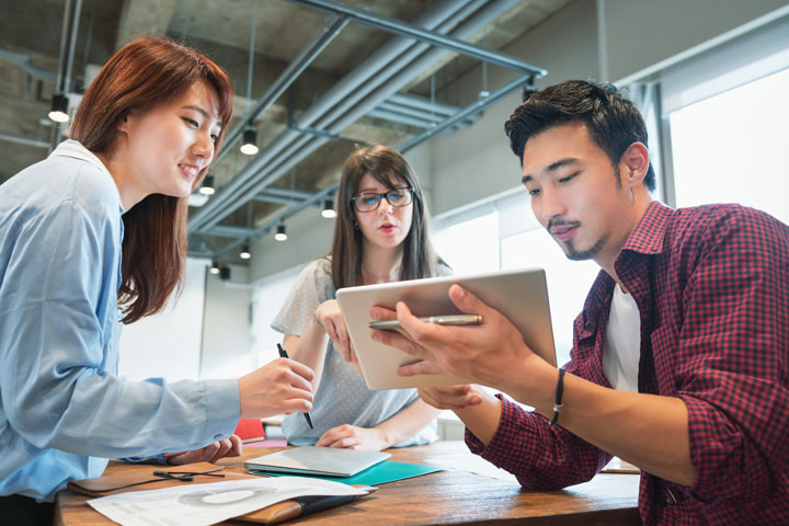 Three students collaborating, with one person holding a tablet and the others looking on and discussing.