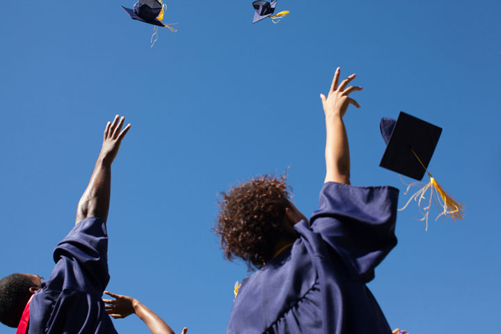 Students wearing their graduation gown, throwing their caps into the sky in celebration after completing and passing their course. The sky is blue with no clouds in sight.