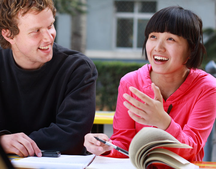 A woman in a pink top smiles and gestures as she talks, holding a book open in front of her. A young man in a black sweatshirt sits next to her, smiling and looking at her while holding a pen.