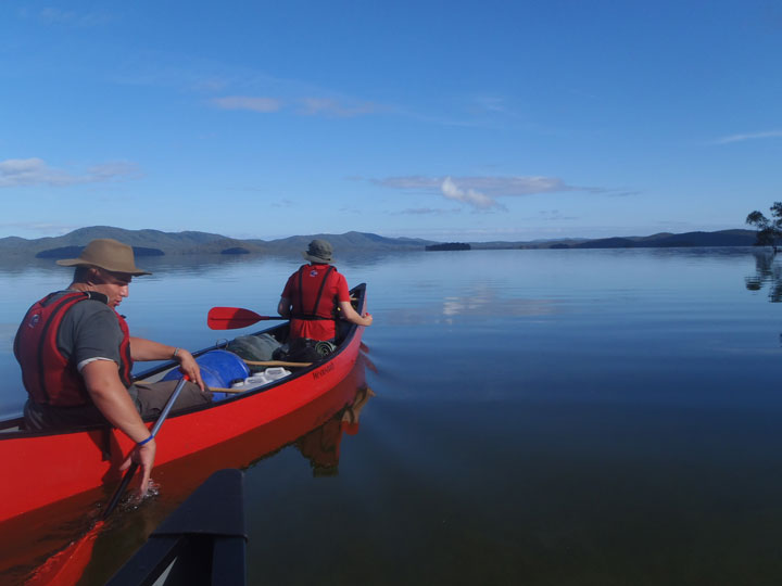 Two people are paddling a red canoe on a calm lake. The person at the front is facing away from the camera, while the person at the back is looking to the side, with one hand in the water. Both are wearing wide-brimmed hats and life jackets. The scene is peaceful and serene, with clear weather and smooth water.
