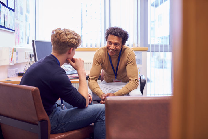 A male student sits in an office with a male counsellor. The student is facing the counsellor, who has curly hair and is smiling warmly. The office setting includes a desk, computer, and various papers and posters on the walls.
