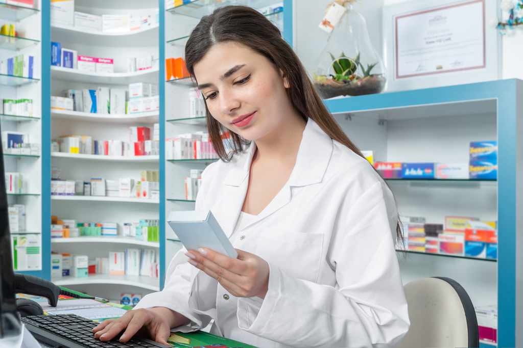 Female pharmacist in a white lab coat examining a product box while working at a computer in a well-stocked pharmacy.