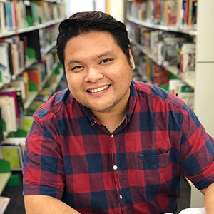 Ron Rocio is sitting in a library smiling at the camera and wearing a black and red check shirt.