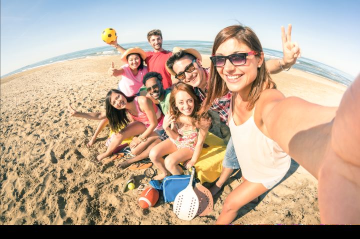 A group of friends are enjoying a day at the beach. They are sitting close together on the sand, smiling and posing for a selfie. The group is diverse and dressed in casual beachwear. The scene is sunny with clear skies, and various beach items like a ball, paddles, and a bag are visible around them. The background features the ocean and a sandy beach, creating a fun and relaxed atmosphere.