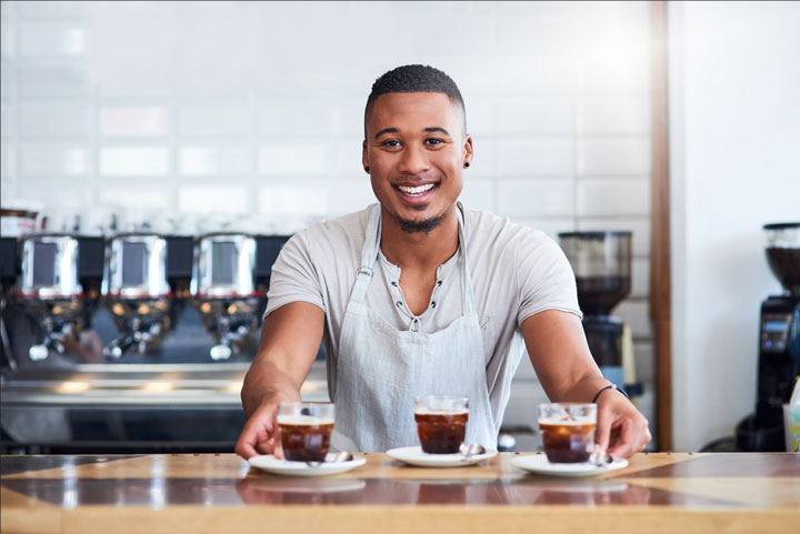 A smiling barista who is a young male is wearing a pastel blue apron over a grey tee shirt, is standing behind a counter, presenting three cups of coffee on saucers. He is smiling at the camera. The background features coffee machines and a bright, clean café environment, creating a warm and inviting atmosphere.
