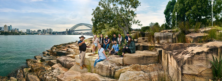 A group of people is gathered on a rocky shoreline overlooking Sydney Harbour. In the background, the Sydney Harbour Bridge and city skyline are visible under a clear sky. The group is engaged in an outdoor educational activity, led by a man wearing a hat and holding a plant. The setting is lush with greenery and the people appear attentive and interested.