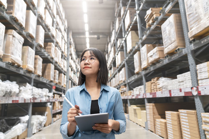 A woman with glasses stands in a large warehouse, holding a tablet and stylus, looking up thoughtfully at the tall shelves stocked with boxes.