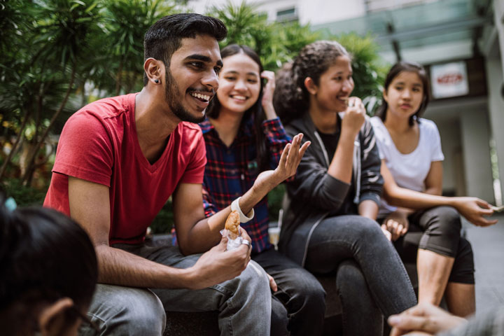 Four students sit on a bench seat, feeling content, relaxed and enjoying the company. The man closest to the camera is gesturing with one hand, holding a pastry in the other and is animatedly speaking to other people outside the camera shot.