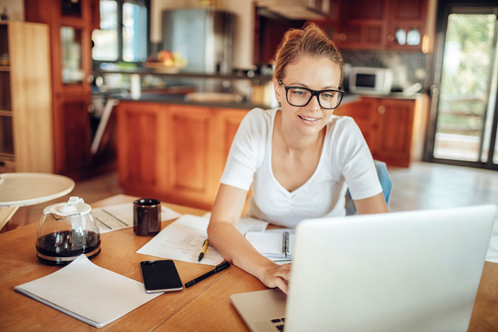 A woman is working at a wooden table in a cosy kitchen. She is wearing glasses and a white shirt, smiling as she looks at her laptop. The table is scattered with papers, a notebook, a pen, and a smartphone.