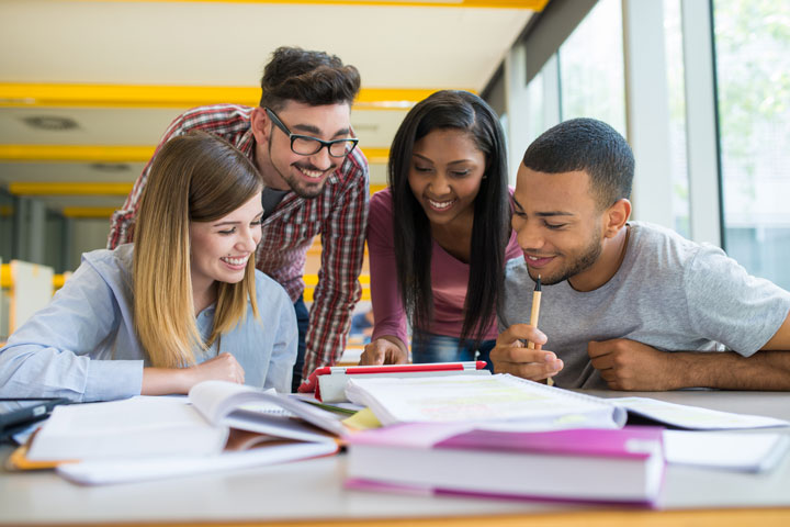 A group of four diverse young adults, two men and two women, are gathered around a table in a bright, modern study area. They are smiling and engaged in collaborative work, looking at a tablet and various open books and notebooks spread out in front of them.