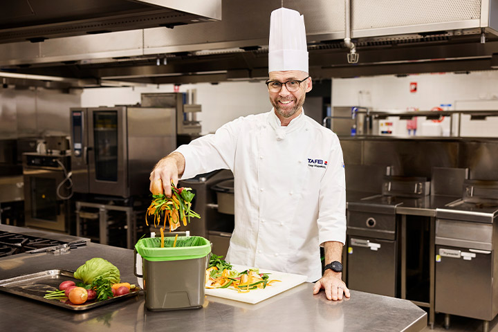Chef wearing a TAFE NSW uniform and toque stands in a commercial training kitchen placing vegetable scraps into a food waste caddy, demonstrating sustainable food waste practices with fresh produce and stainless steel kitchen equipment visible in the background.