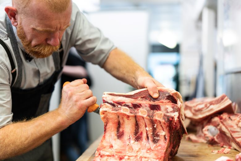 Close-up of a butcher in an apron, intently carving a rack of ribs on a wooden butcher’s block, with a focused and precise expression.