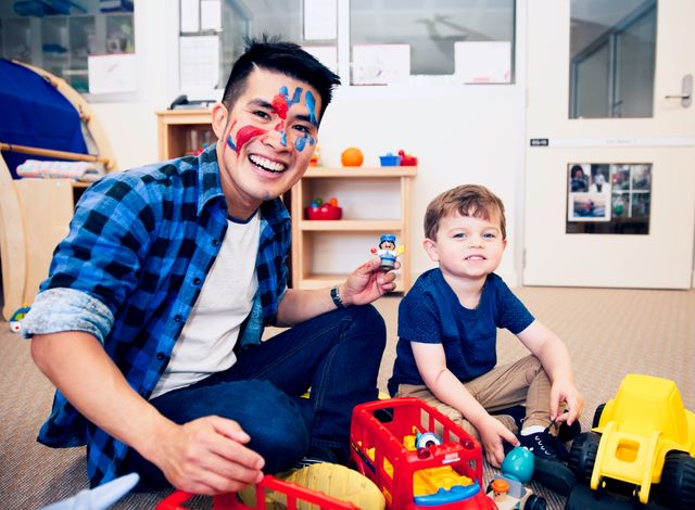 A smiling man with face paint and a blue plaid shirt sits on a playroom floor, holding a small toy figure. Beside him, a young child with short brown hair, wearing a navy blue shirt, plays with colourful toy vehicles.