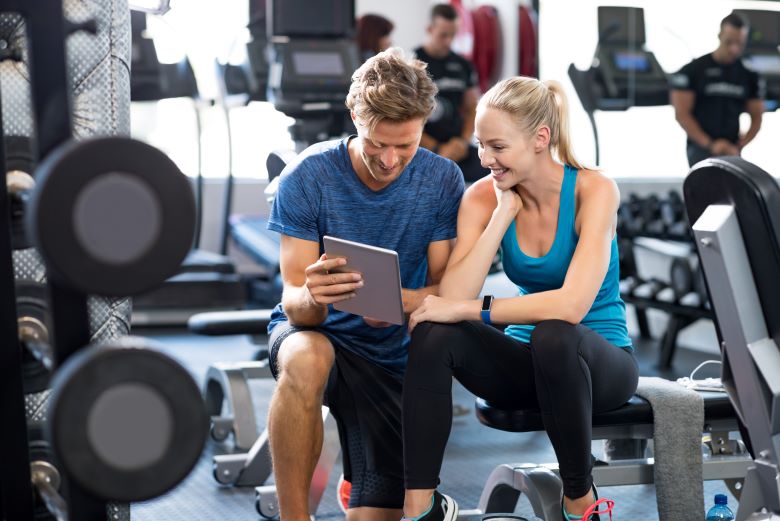 A man and a woman sit on the gym equipment in a fitness centre, smiling as they look at a tablet. The man wears a blue T-shirt and shorts, while the woman wears athletic clothing. Exercise machines and weights are visible in the background.