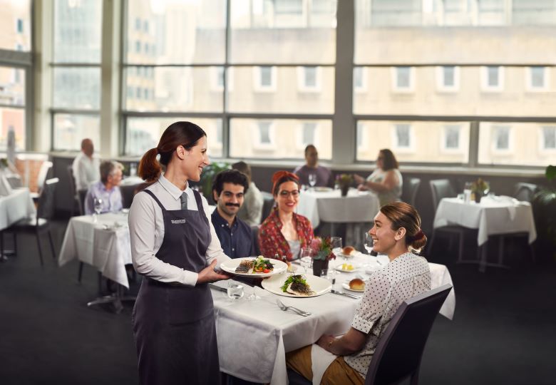 A woman in a tie and apron hands a plate of food to a table of smiling customers. They are seated in a fine dining setting with a view of a city building. In the background, other patrons enjoy their meals while seated at white table-clothed tables. 