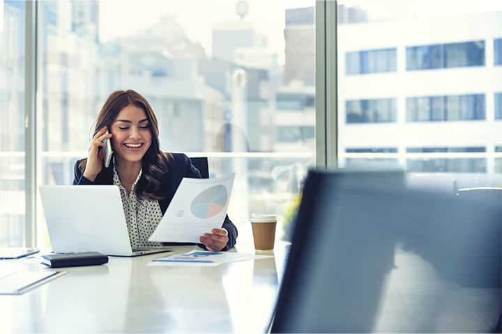 A smiling woman is working in a modern, well-lit office. She is talking on the phone and looking at a document.