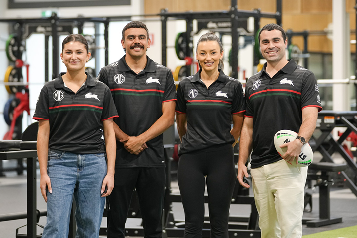 Group of four Rabbitohs Institute of Sport students in matching black polo shirts with the Rabbitohs logo, standing in a gym with exercise equipment.