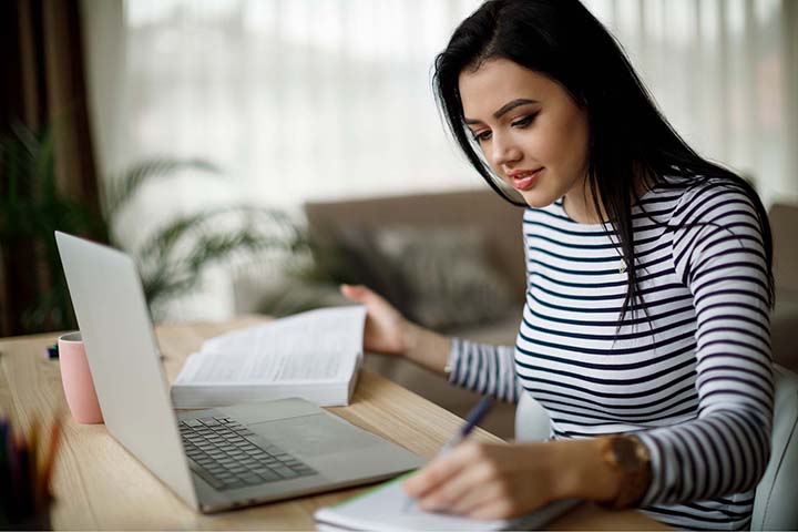 A young lady working from home at a table with her laptop open, writing notes in a notepad with her left hand and holding an open book in her right hand.