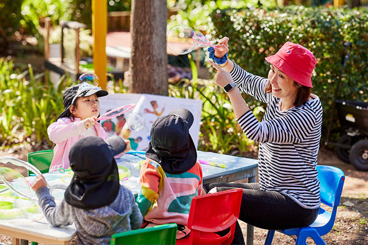 Adult supervising a bubble-blowing activity with young children seated around a table in an outdoor play area, promoting child-safe engagement.