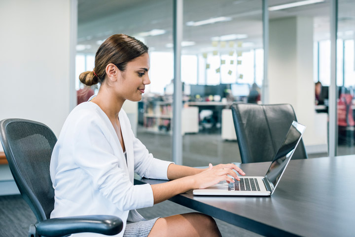 Woman working on a laptop at a desk in an open-plan office, with glass walls and office furniture in the background.