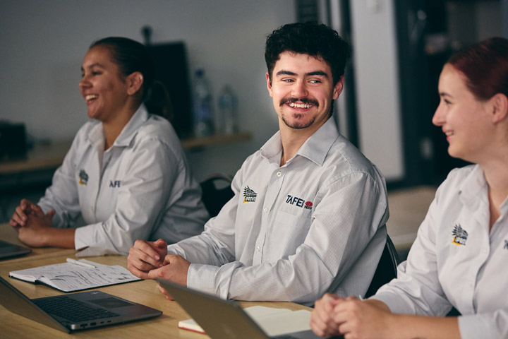 Three IPROWD students in a classroom setting wearing white shirts with TAFE NSW and IPROWD logos, seated at a table with laptops, notebooks, and papers, with shelves and water bottles in the background.