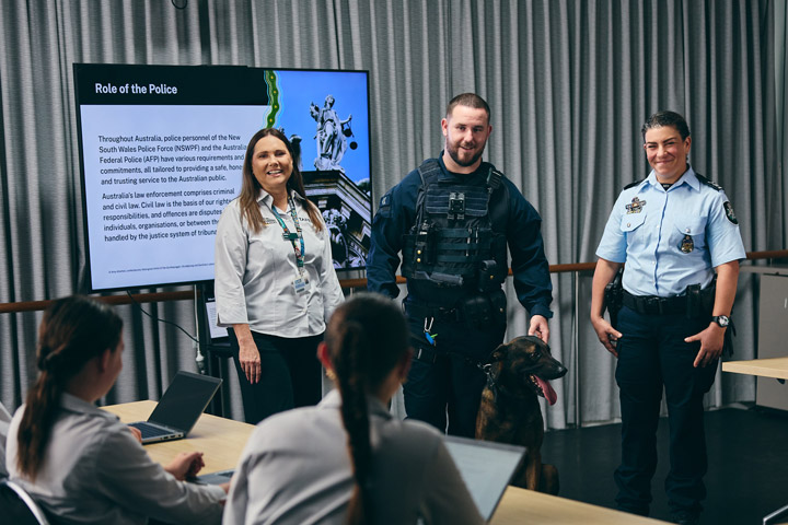 ɫֱIPROWD classroom session where two NSW Police Officers, one holding a dog, and a woman are presenting to students seated at desks with laptops. A screen behind them displays the title 'Role of the Police' along with information about law enforcement responsibilities in Australia.